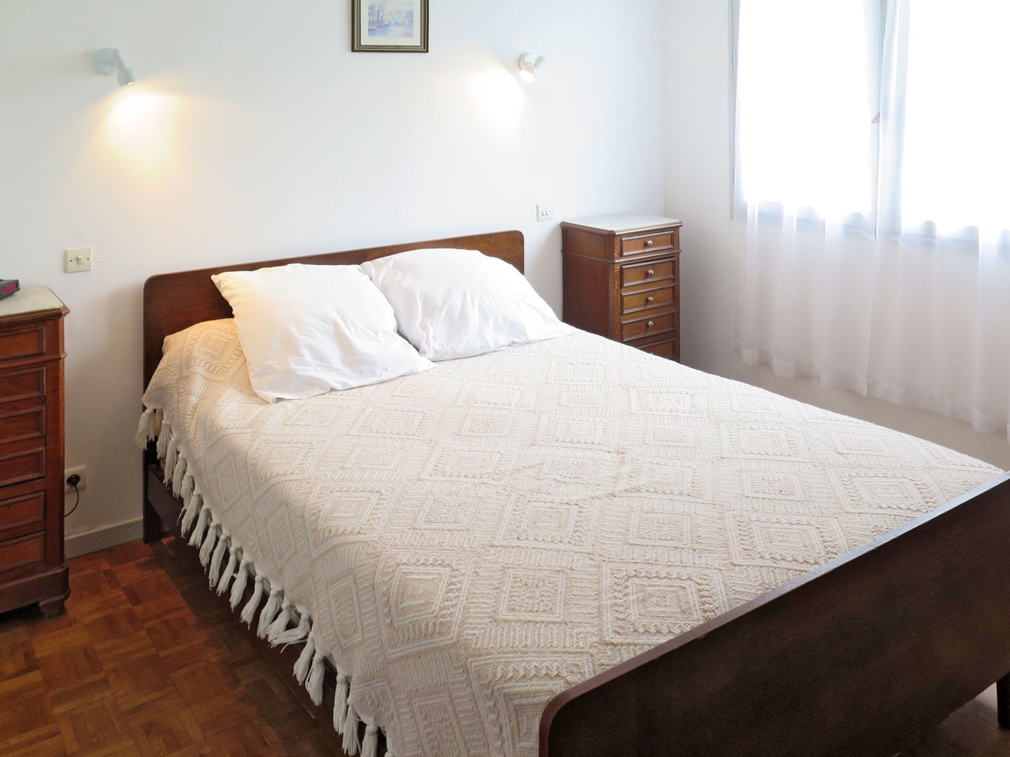 A neatly made bed with a patterned bedspread and pillows, a wooden nightstand with a lamp, and a small dresser with drawers.