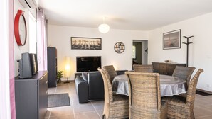 Ein Wohnzimmer mit Fernseher, Sofa, Esstisch mit Stühlen und Bildern an den Wänden.