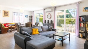 Ein modernes Wohnzimmer mit einer grauen Eckcouch, einem Couchtisch und einem Bücherregal. Es gibt eine Essecke mit Stühlen und einem Tisch, sowie ein großes Fenster mit Blick ins Freie.