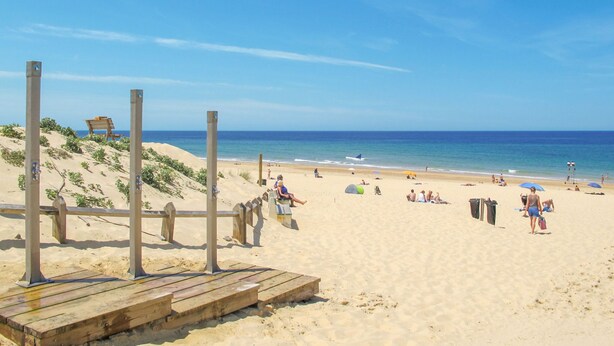 Ein sandiger Strand mit Holzgebäuden, blauem Himmel und dem Meer im Hintergrund.