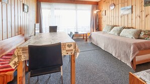 A cozy room with a wooden interior, a bed with patterned bedding, a dining table with chairs, and a view through large windows. - Greppon 306 by Interhome (Thyon-Les Collons)