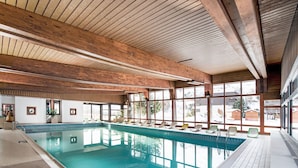Une piscine intérieure avec des poutres en bois, de grandes baies vitrées et des chaises longues disposées le long du bord de la piscine.
