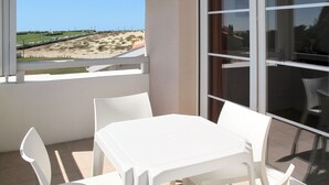 A balcony with a white table and chairs, a glass door, and a view of a beach and parking lot.