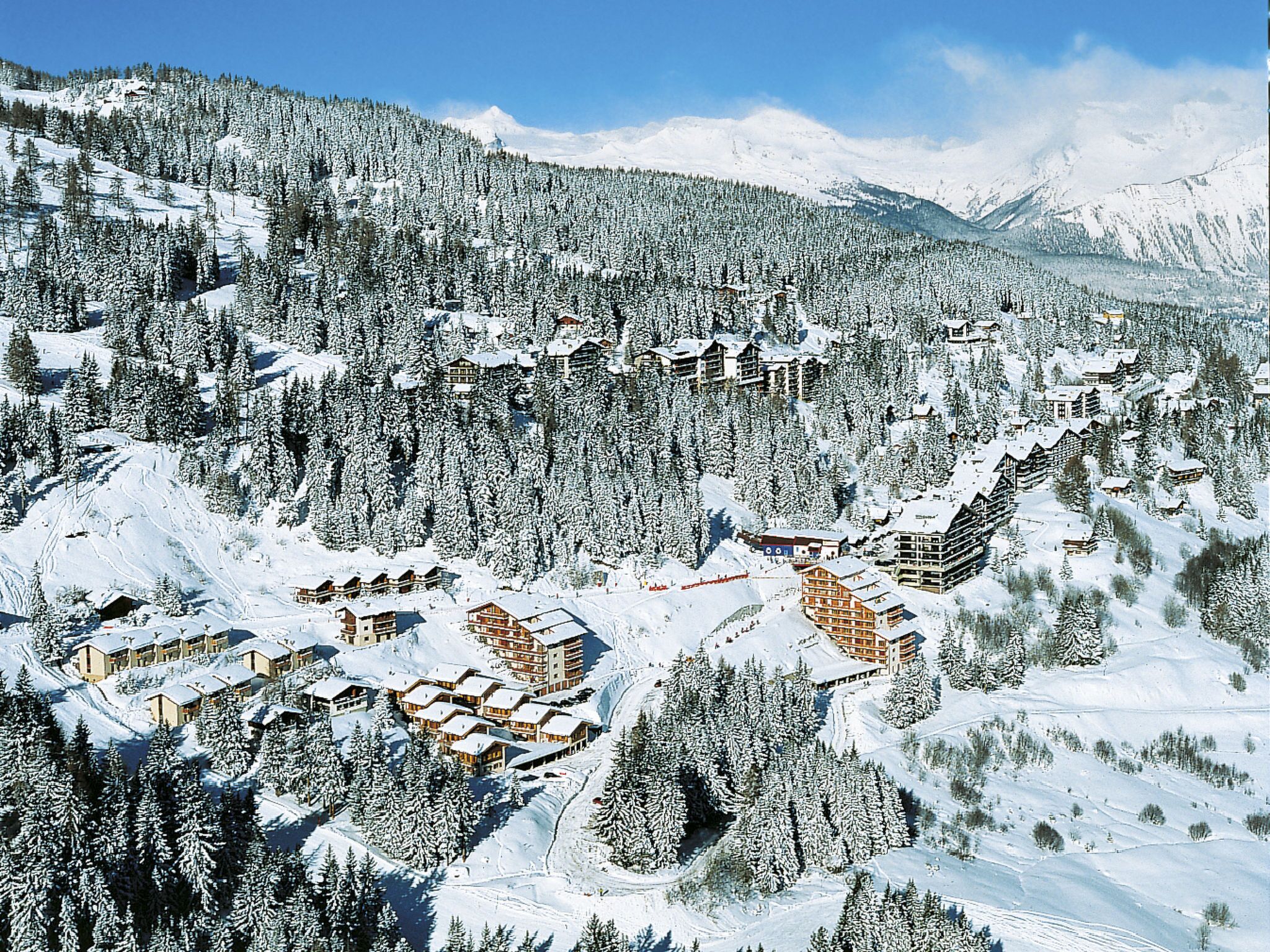A snow-covered mountain resort with multiple chalet-style buildings, dense forest, and a clear sky.