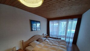 A bedroom with a wooden bed, a bedside table, a lamp, a window with sheer curtains, and a framed picture on the wall. - Essert 101 by Interhome (Thyon-Les Collons)
