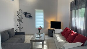 A modern living room with a grey sofa, a coffee table with a vase, a flat-screen TV on a stand, and a window with blinds. - Le Moïsan by Interhome (Messanges)