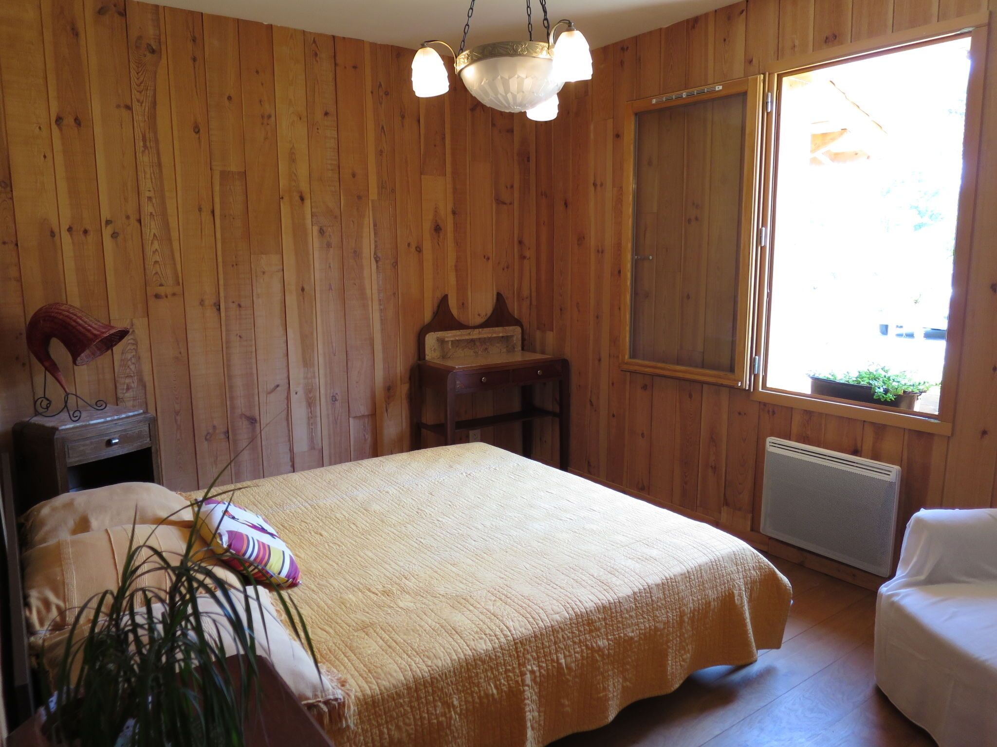 A wooden-paneled room with a bed, a small table, a chair, a radiator, and a window with a view.