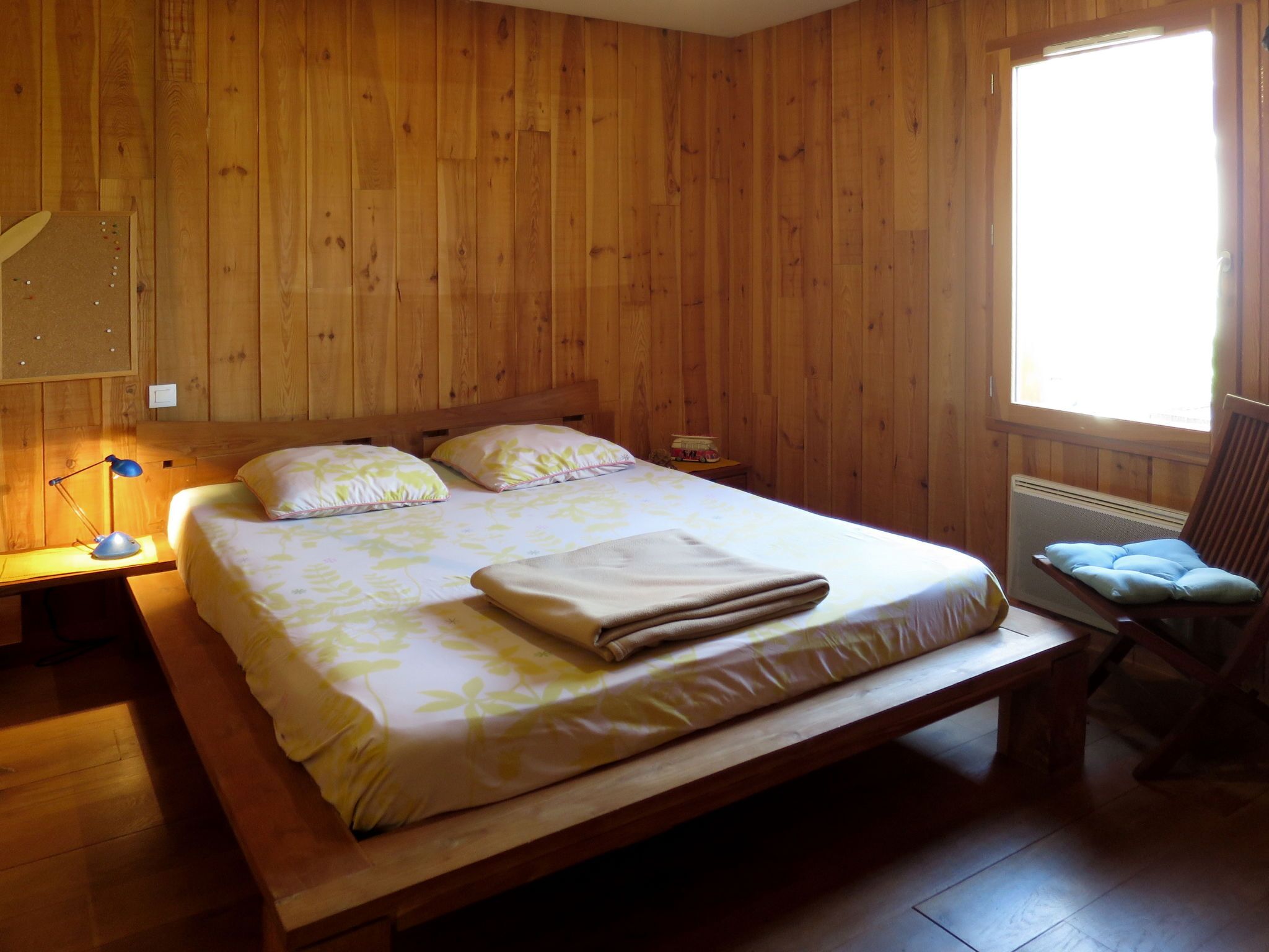 A wooden-paneled room with a bed, a chair, a nightstand, and a window.