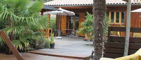 Una terraza de madera con sillones de descanso y una mesa, rodeada de macetas con plantas y una palmera.