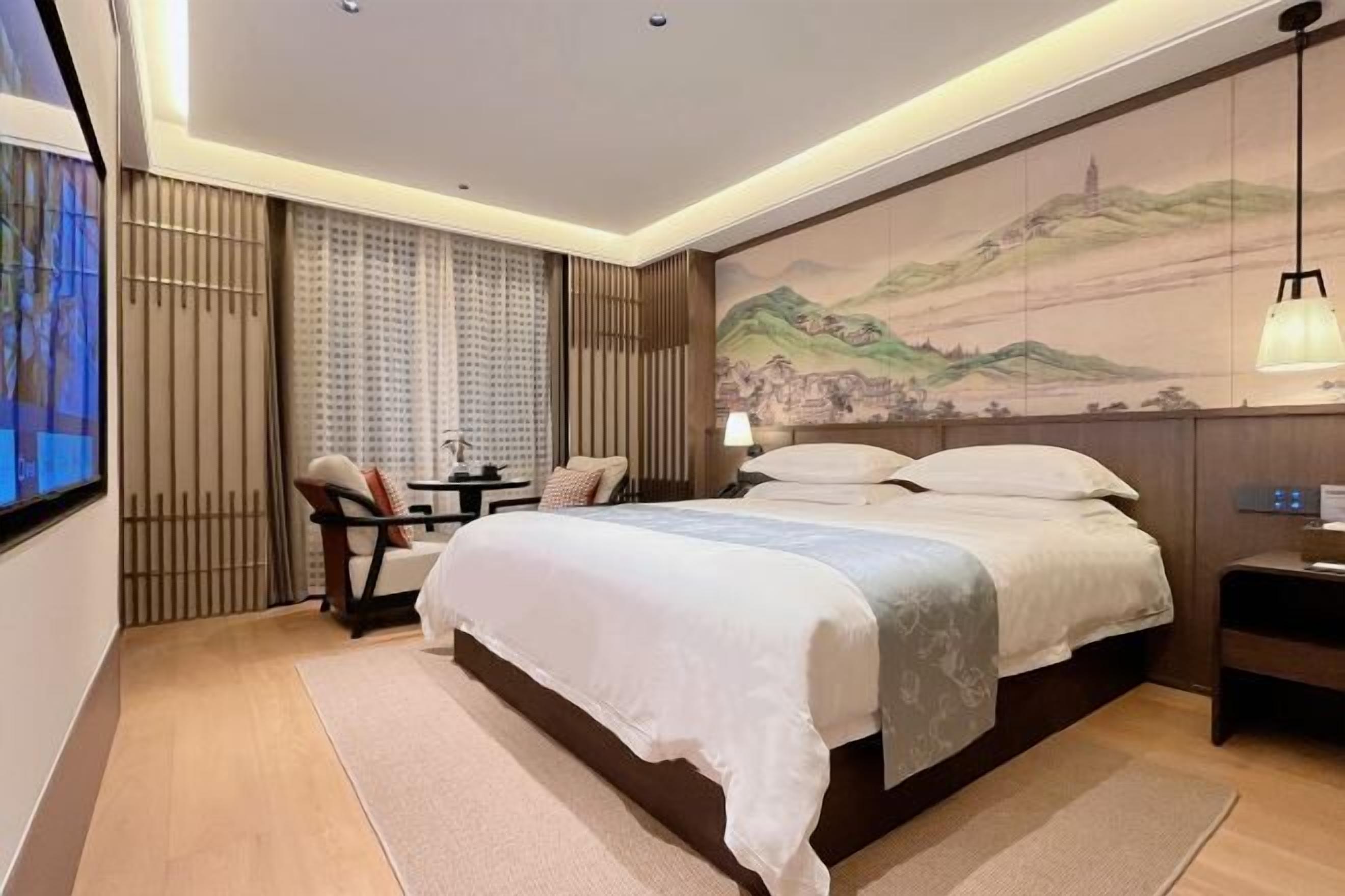 A hotel room with a large bed, a desk with a chair, a small table, and a wall-mounted lamp.