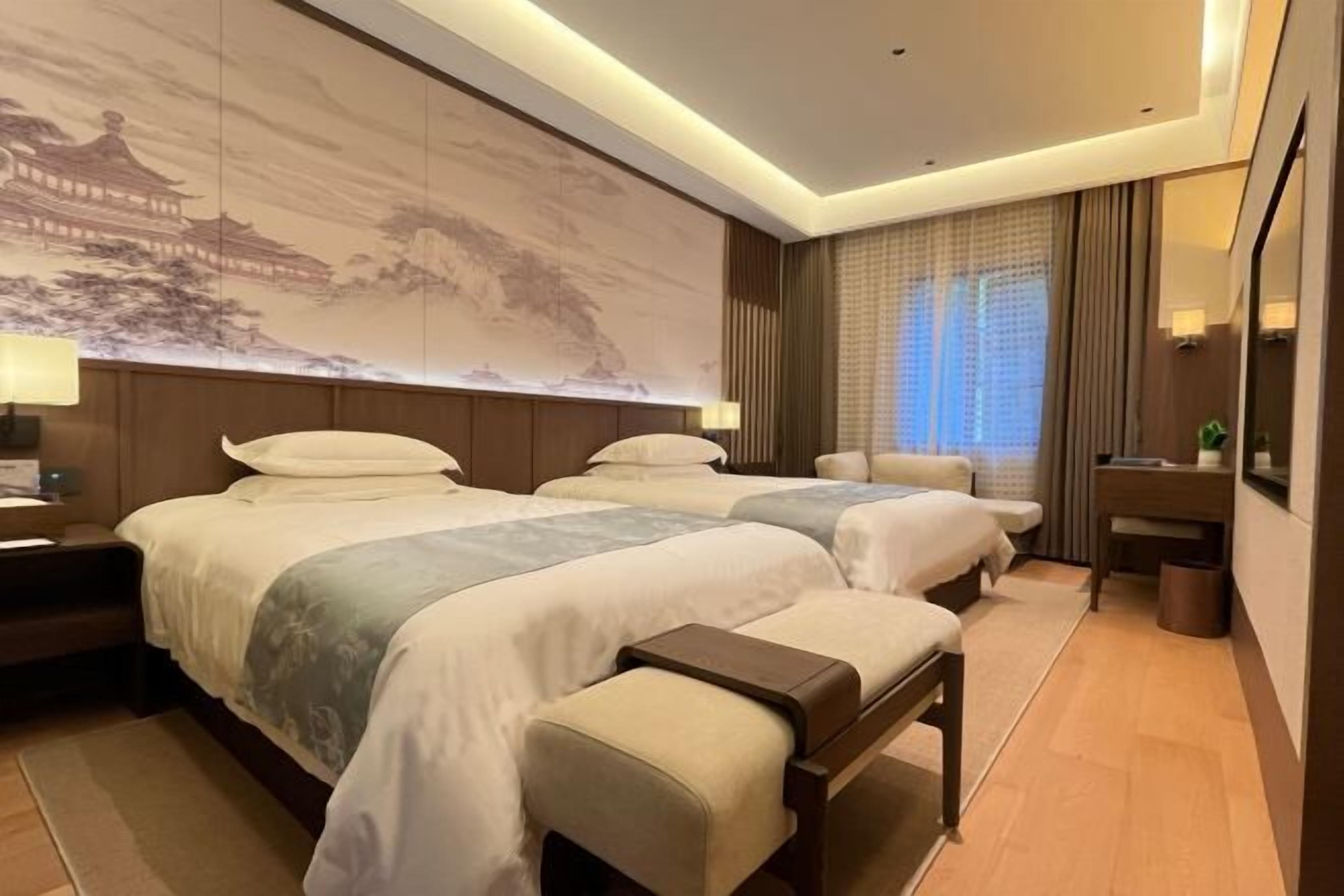 A hotel room with two beds, a large wall mural, a desk, and a window with curtains.