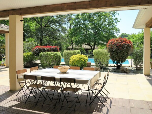 An outdoor patio with a table and chairs, surrounded by well-maintained hedges and trees, with a pool in the background.