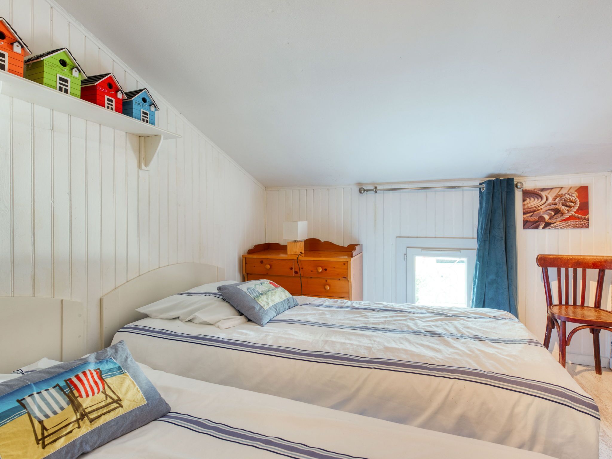 Un dormitorio con dos camas, un aparador de madera, una silla y una estantería con casas de madera de colores.