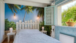 A bedroom with a large bed, a mural of a tropical scene, a window with a view of greenery, and a small table with a lamp. - Babelle by Interhome (Gaillan-en-Médoc)