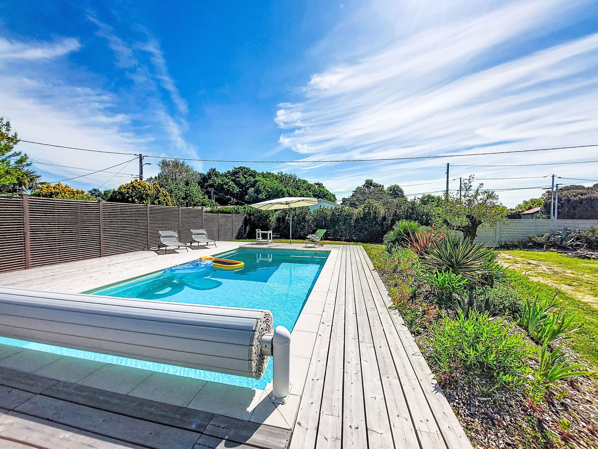 A swimming pool with a wooden deck, surrounded by greenery and a clear sky.