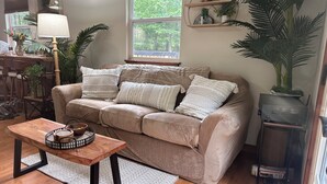A cozy living room with a beige sofa, a wooden coffee table, and a large window with a view of greenery. - Luxury Cabin, fire pit, BBQ, WiFi, forest view, river access (Richland)