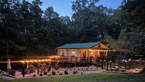 A wooden cabin with a deck, surrounded by trees and a pond, featuring string lights and outdoor seating. - Luxury Cabin, fire pit, BBQ, WiFi, forest view, river access (Richland)