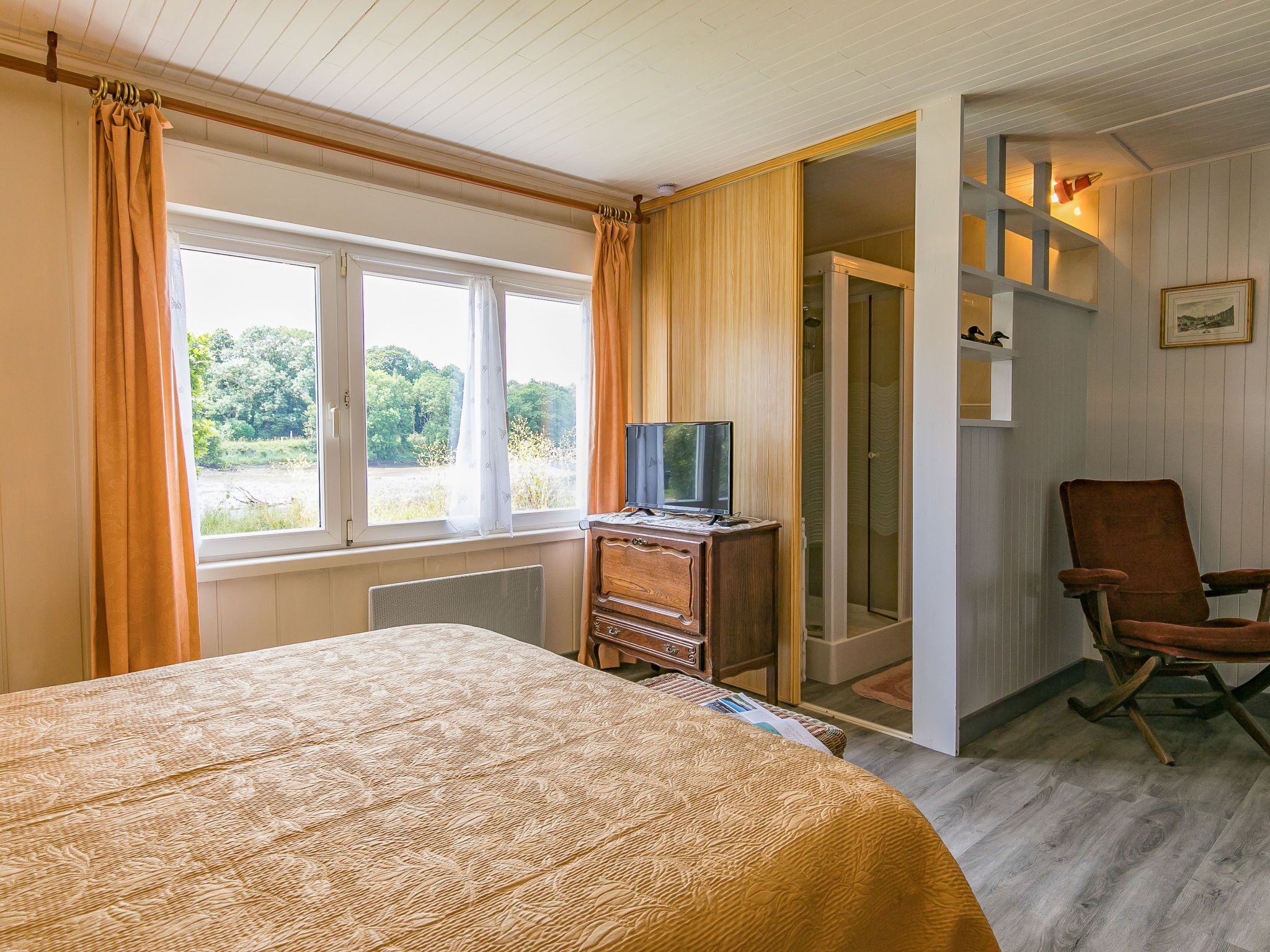Une chambre à coucher avec un lit, un téléviseur posé sur une commode en bois, une chaise et une vue sur l’extérieur par une fenêtre.