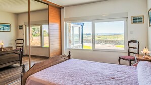 A bedroom with a large bed, a wooden headboard, a mirror, a window with a view of the countryside, and a small table with a lamp. - Blaues Meer by Interhome (Plouarzel)