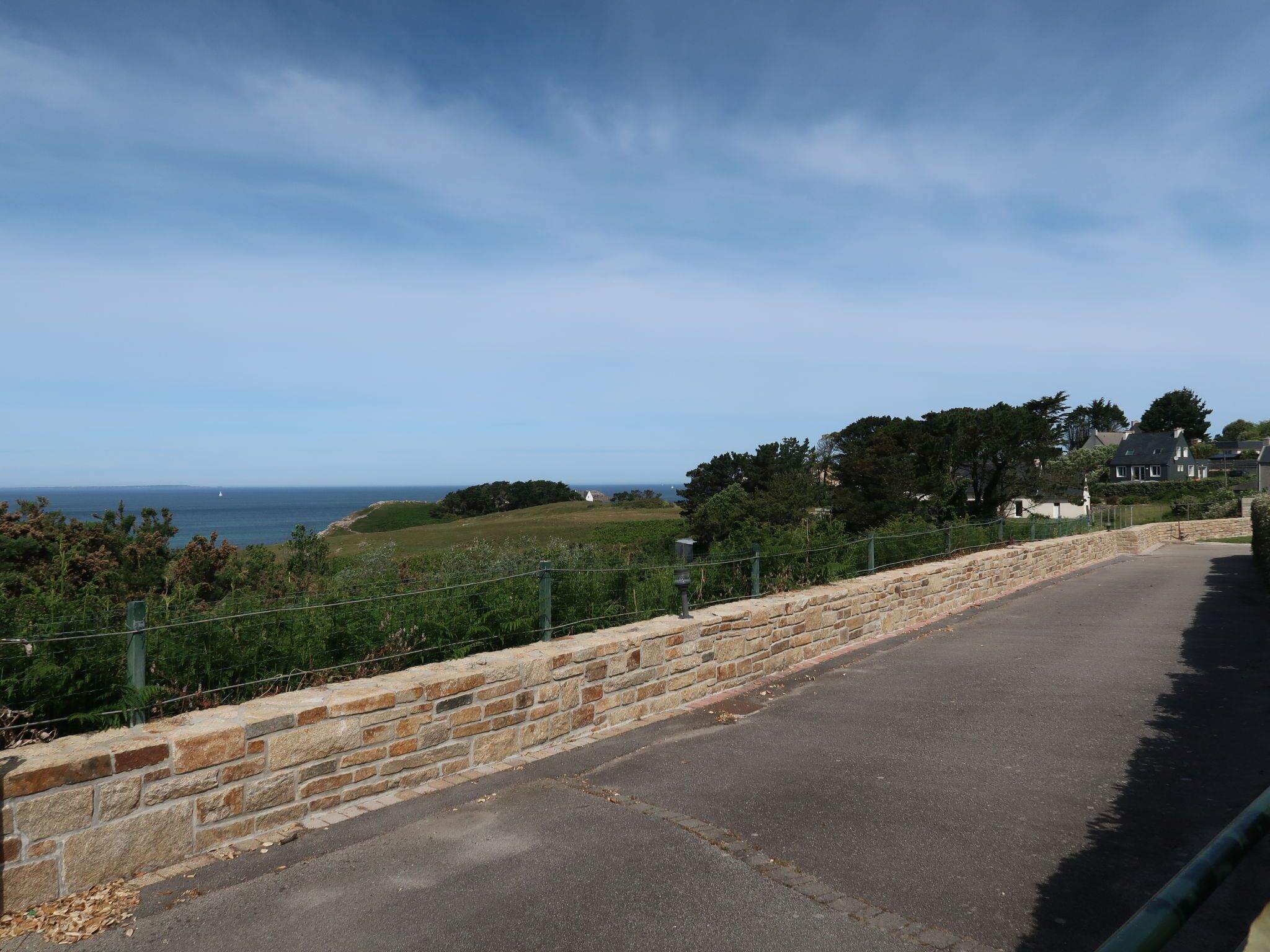 A coastal road with a stone wall, greenery, and houses in the background.