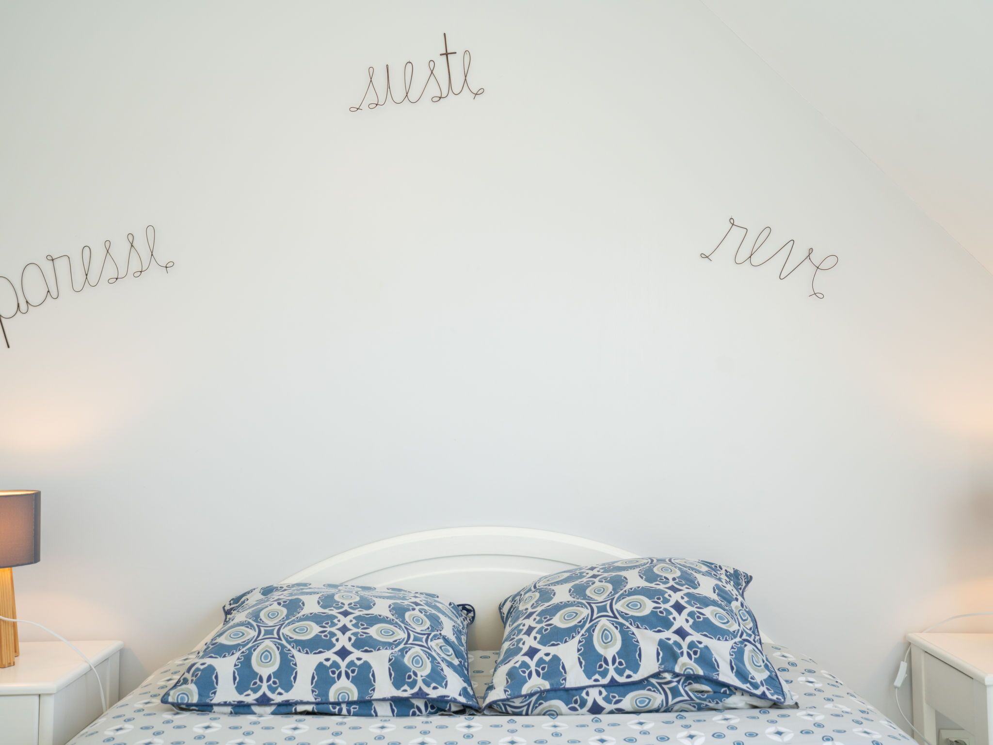 Un dormitorio con cabecera blanca, dos almohadas estampadas y una mesita de noche con lámpara.