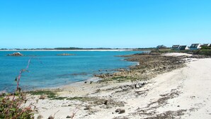 A coastal beach with rocky shoreline, clear blue water, and houses in the background.