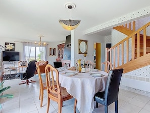 Un coin repas avec une table dressée pour quatre personnes, entouré de chaises en bois, d’un lustre et d’un escalier orné d’une rampe à motifs.