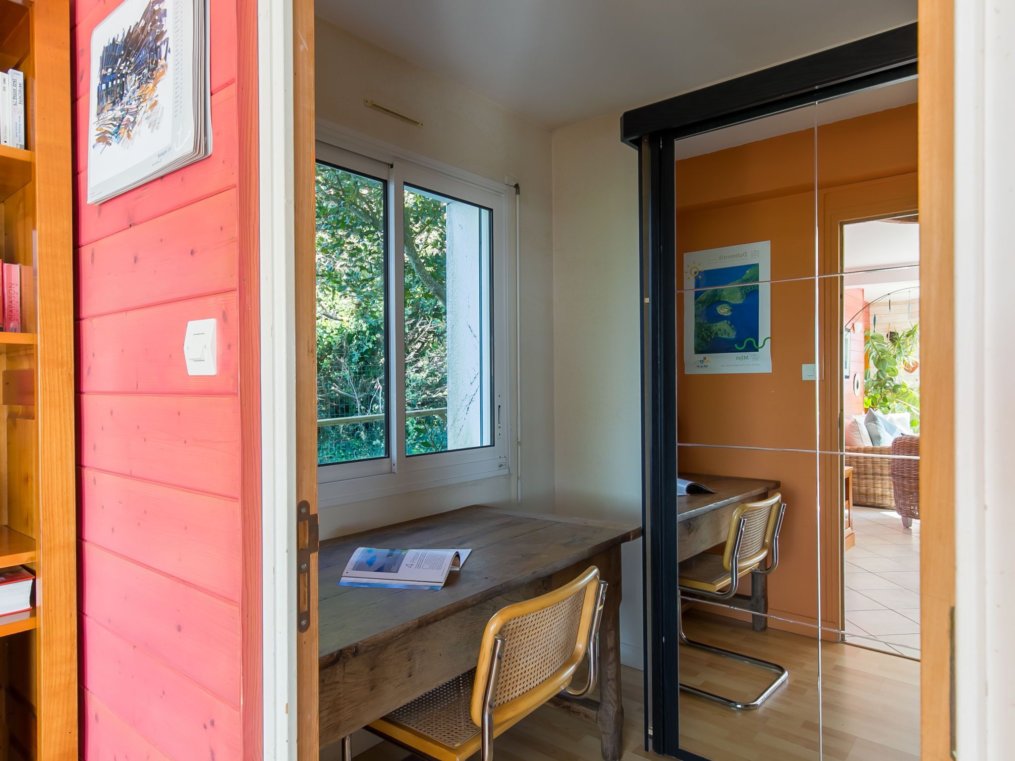 Una habitación con un escritorio de madera, dos sillas, un ventanal con vistas a la vegetación y una puerta que da a un espacio exterior con una silla de mimbre.
