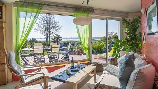 Una habitación luminosa con vistas a un jardín y un cuerpo de agua. Cuenta con una mesa de comedor, sillas, un sofá y un ventanal con cortinas.