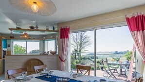 Un comedor con mesa preparada para cuatro personas, ventanas grandes con cortinas y vista al campo.