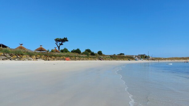 Une plage avec un ciel dégagé, un rivage sablonneux et quelques huttes aux toits de chaume.