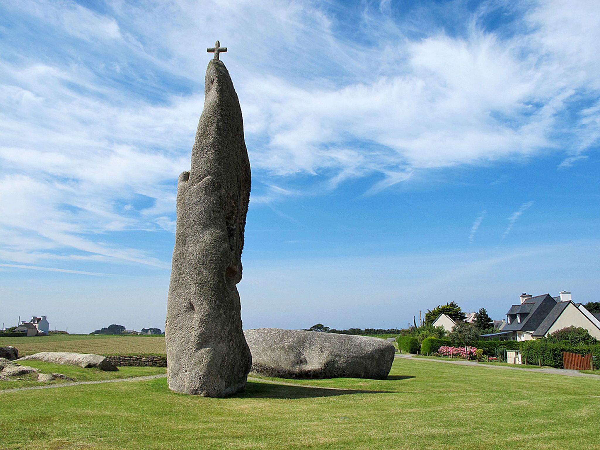 A large standing stone with a cross on top, set in a grassy field with houses in the background.