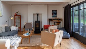 Un salon spacieux avec un plafond haut, un poêle à bois, un canapé confortable, une table basse et une grande fenêtre donnant sur l’extérieur.