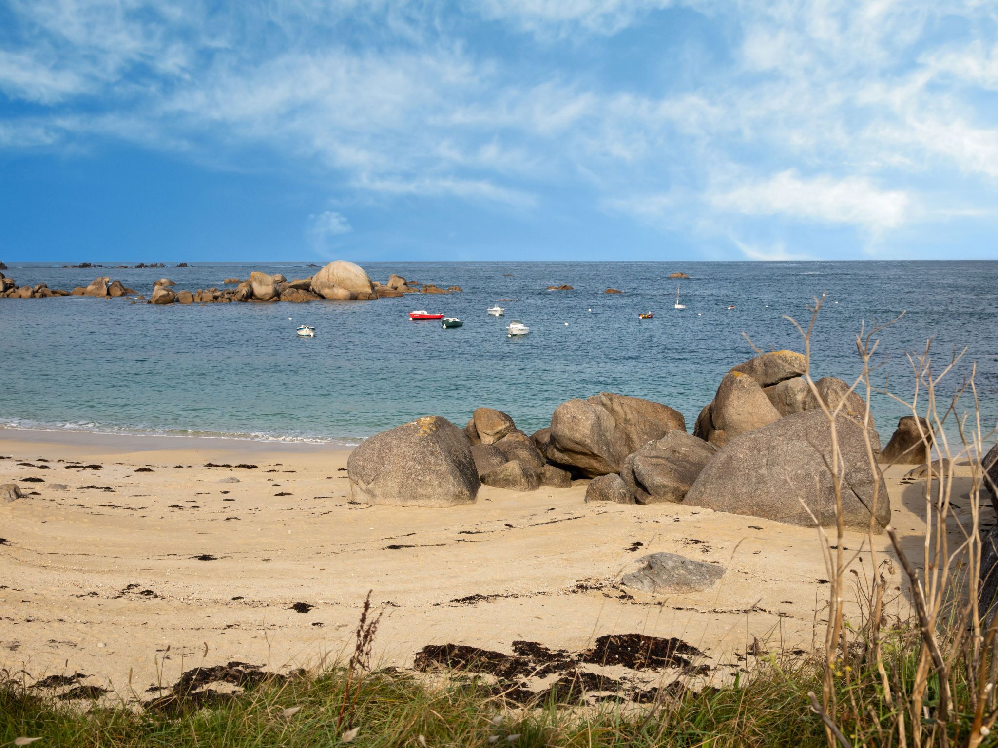 Una playa con rocas grandes, mar tranquilo y barcos.