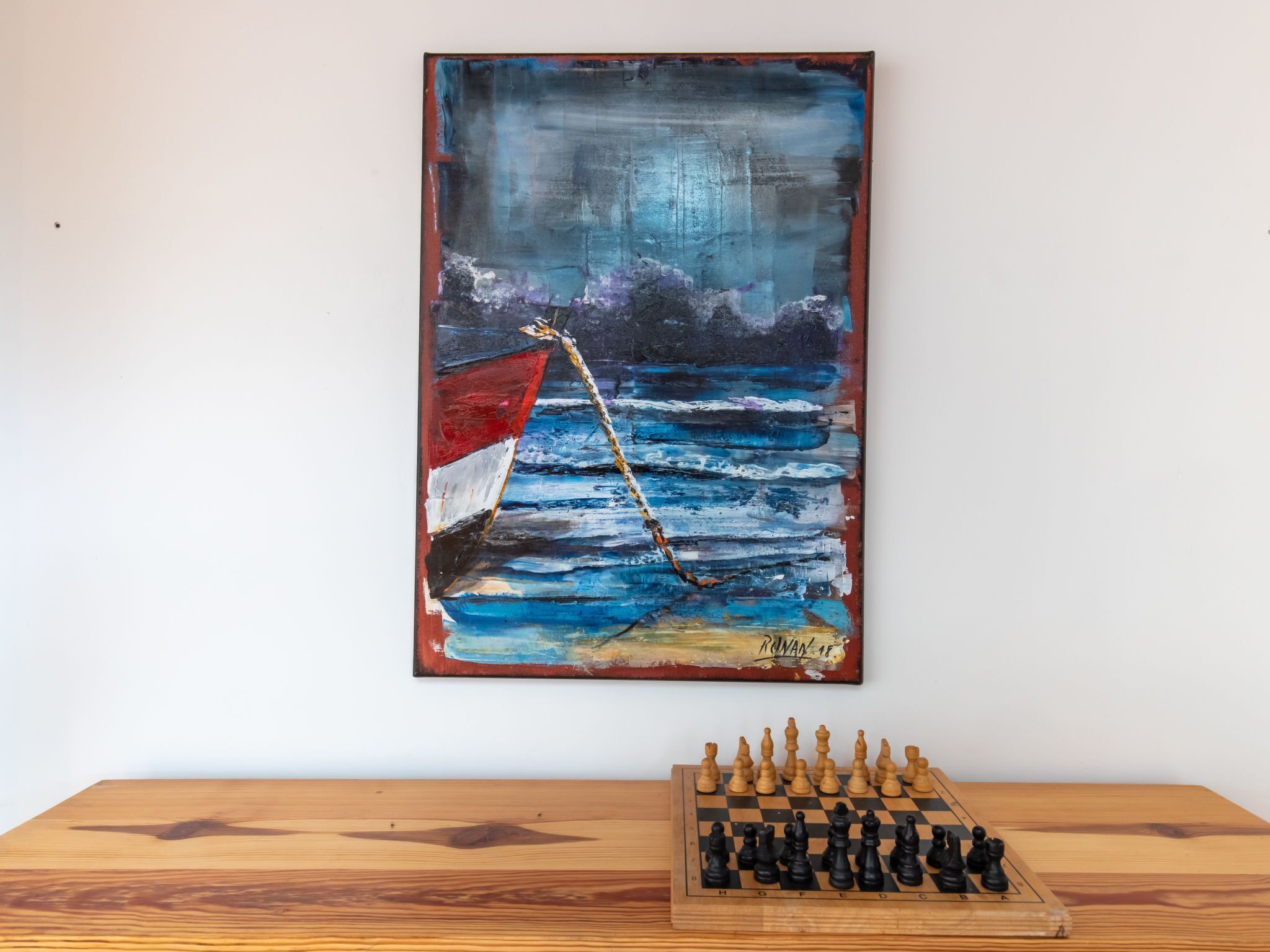 A wooden table with a chessboard and a painting of a boat on a blue sea.