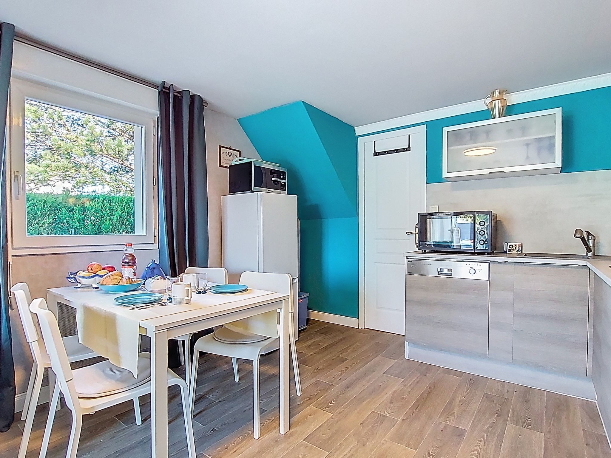 Cozinha compacta com micro-ondas, frigorífico e lava-loiça. Uma mesa de jantar com cadeiras está colocada perto de uma janela com cortinas.