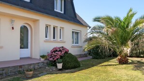 Une maison à deux étages, avec un toit sombre, des murs clairs et une porte d’entrée. Il y a trois fenêtres au deuxième étage et, dans le jardin, un grand pot de fleurs contenant une plante aux fleurs roses.