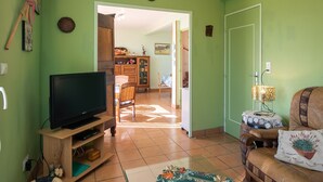 Een woonkamer met een televisie op een houten standaard, een bruine leren bank, een kamerkleed met een patroon, en een eethoek met een houten kast en rieten stoelen.