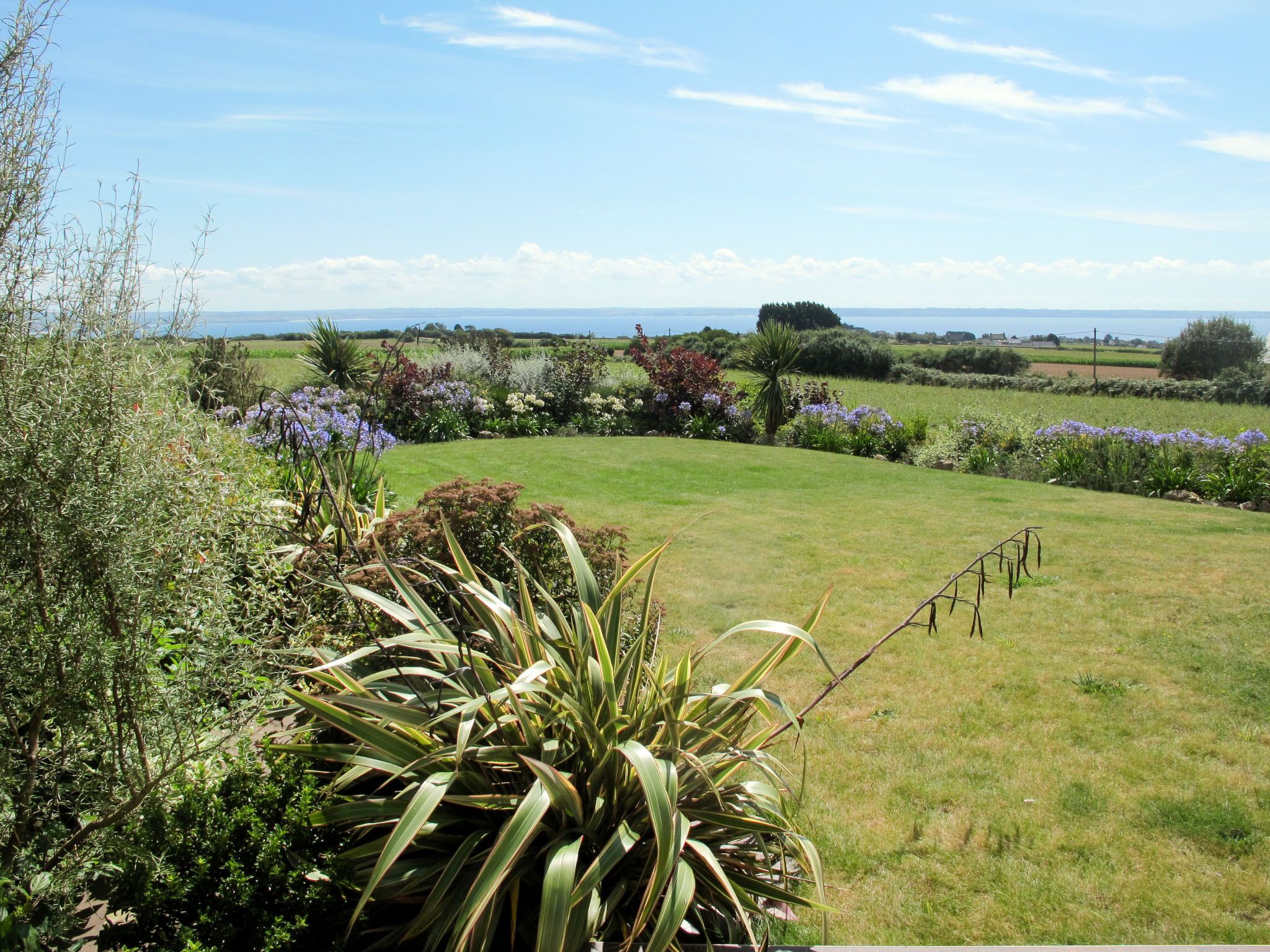 Un jardin bien entretenu, avec une grande variété de plantes et de fleurs, une pelouse verdoyante et une vue sur la mer au loin.