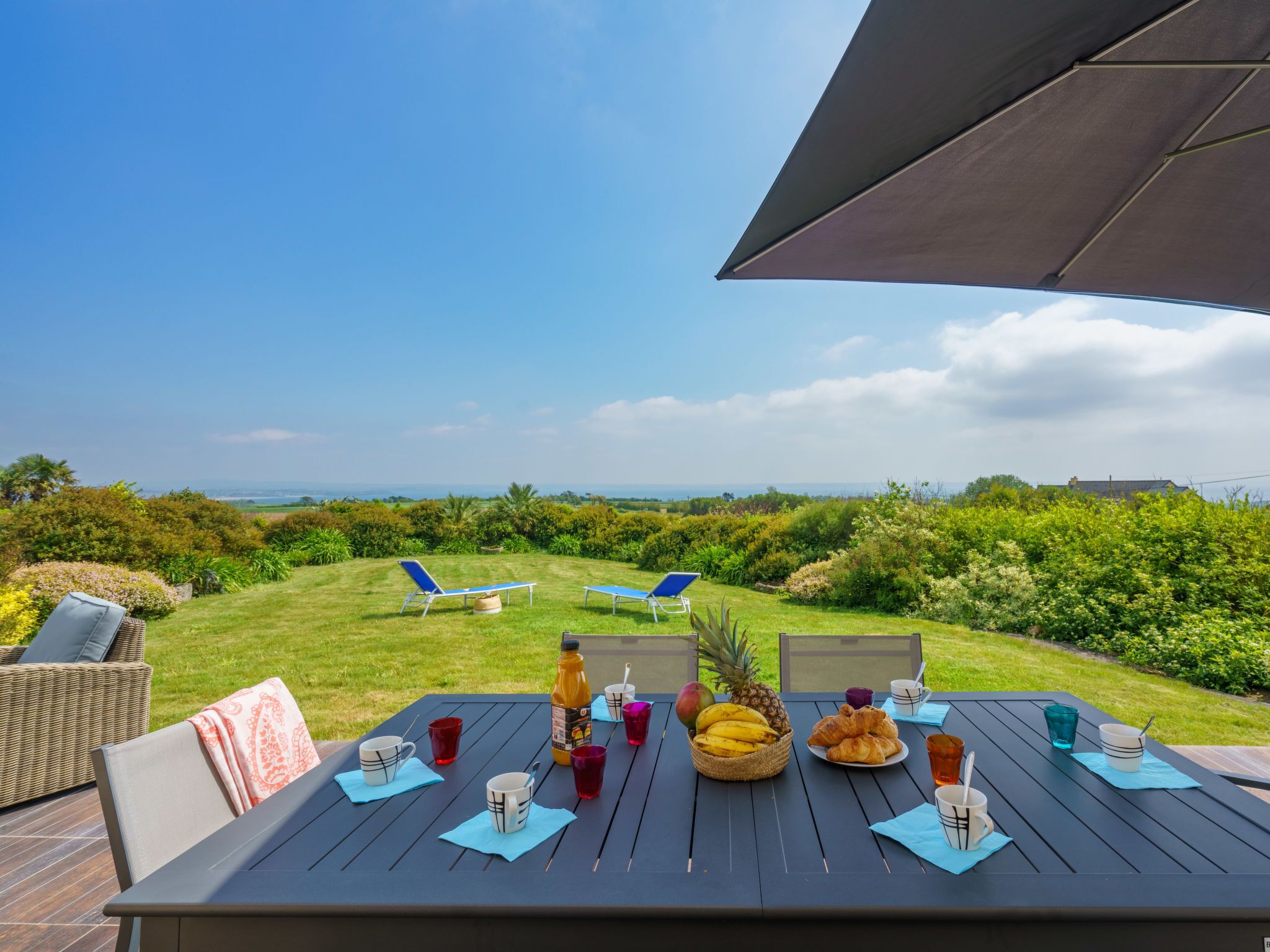 Un espacio exterior para comer con una mesa preparada para dos, que ofrece vistas al océano y abundante vegetación.
