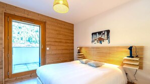 Une chambre avec un grand lit, des murs en bois, une fenêtre donnant sur une forêt et une étagère fixée au mur avec une lampe.