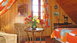 Una habitación acogedora con cama, una silla de mimbre, una mesita con un florero y una pared de madera con un espejo y un cuadro.