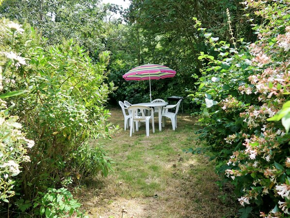 A garden patio with a table and chairs, surrounded by lush greenery and flowering plants.