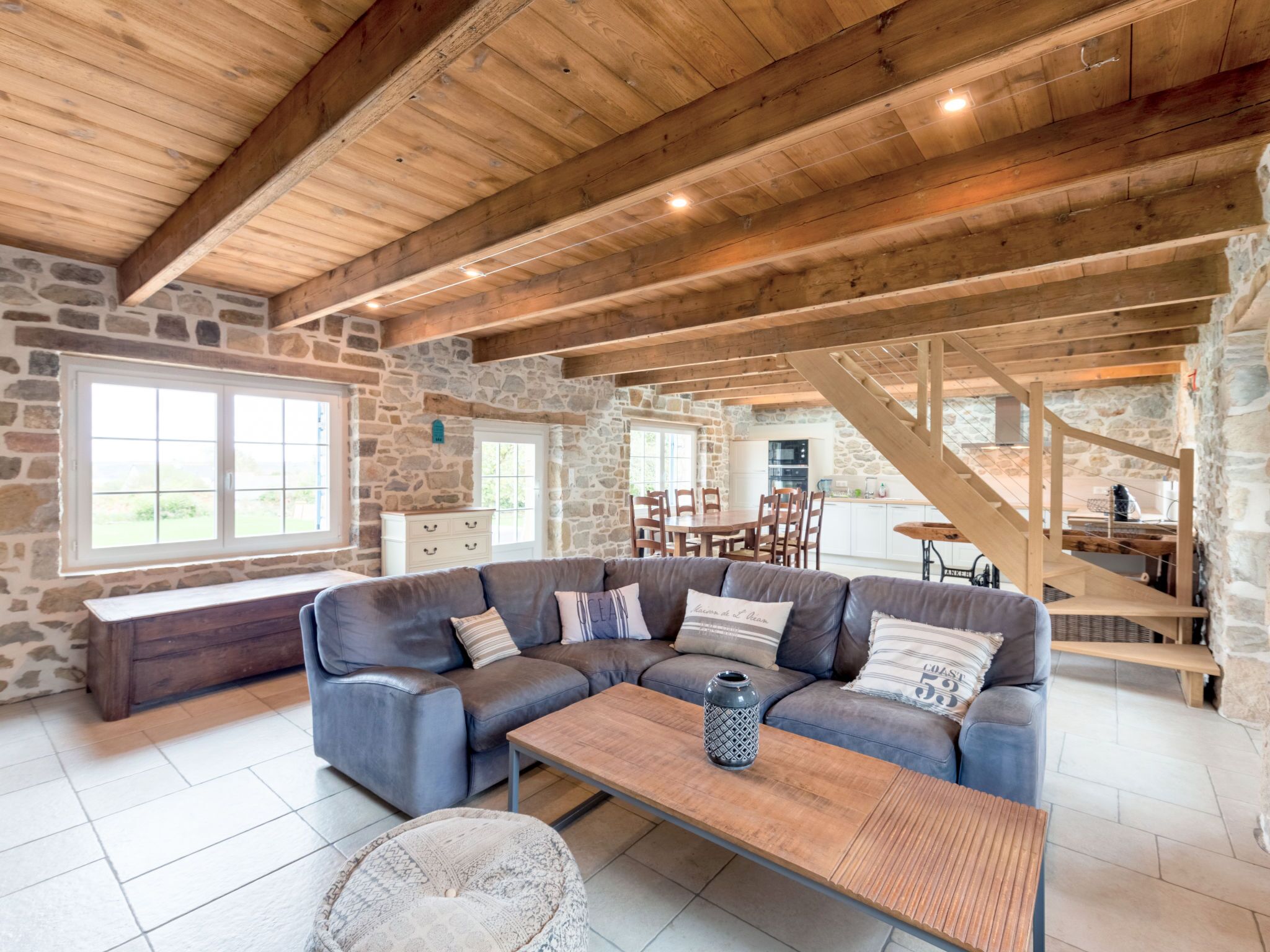 Ein großzügiger Wohnbereich mit Steinwand, Holzbalkendecke, einem Ecksofa und einem Essbereich mit Treppe.