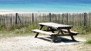 Een houten picknicktafel op een zanderig strand, met een houten hek op de achtergrond en de oceaan in de verte.
