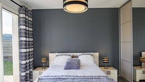A modern bedroom with a large bed, two bedside tables with lamps, a ceiling light fixture, and a view through a large window.