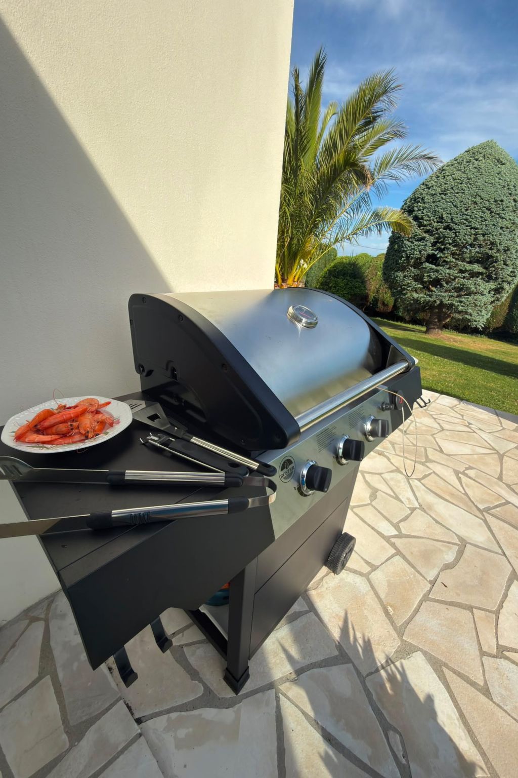 Un barbecue d’extérieur avec une assiette de crevettes cuites, une pince et une brosse pour nettoyer le gril.
