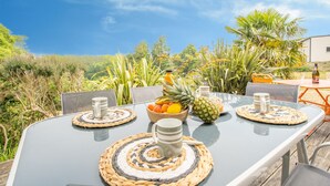 Un área de comedor al aire libre con una mesa preparada para cuatro personas, rodeada de sillas, con vistas a exuberante follaje y un cielo azul despejado.