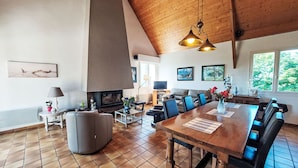 A spacious living area with a high ceiling, a fireplace, a dining table with chairs, a sofa, and a television. - Les Courlis by Interhome (Trévou-Tréguignec)