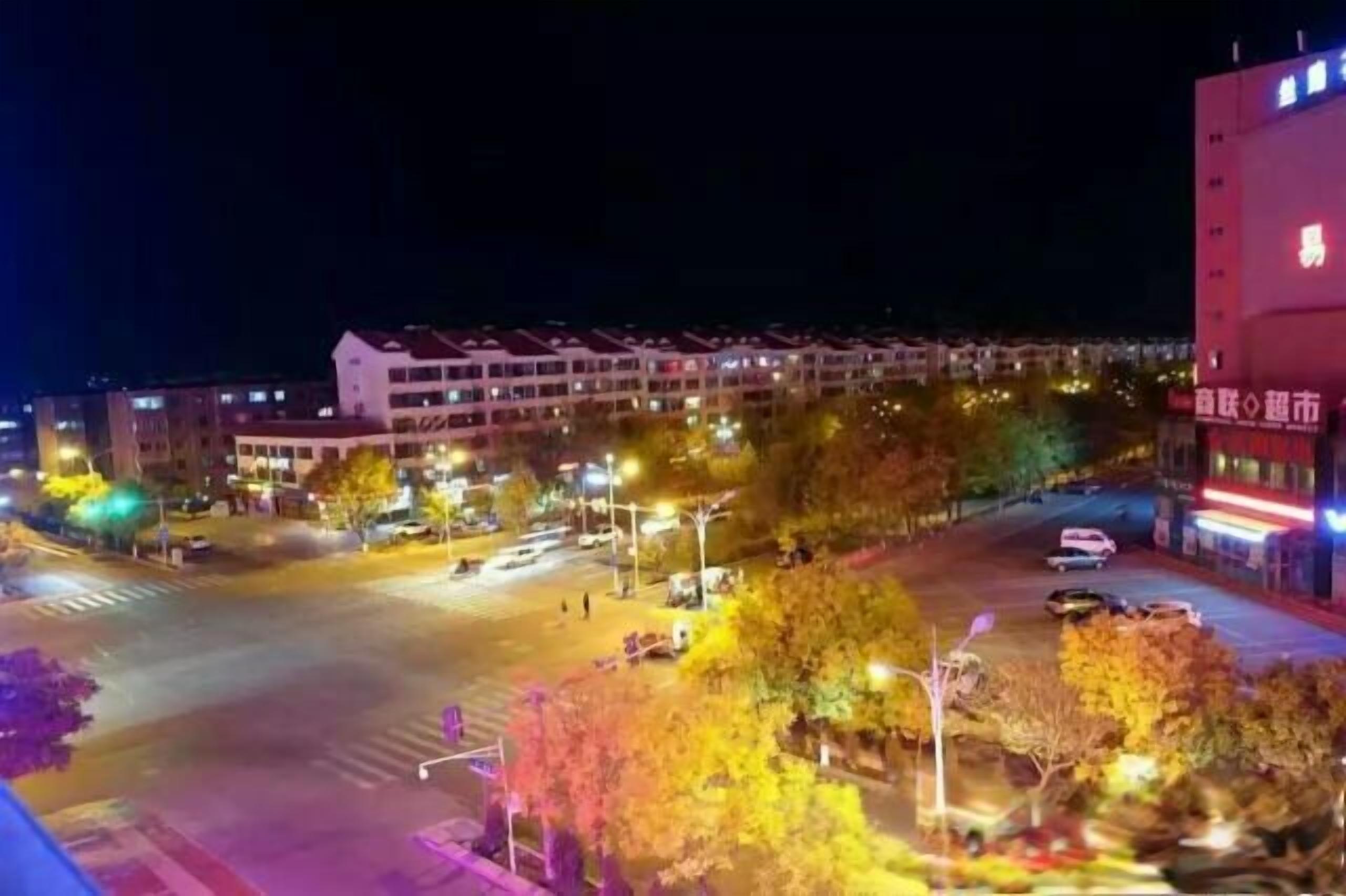 Vista noturna de um cruzamento urbano com edifícios, postes de luz e veículos.